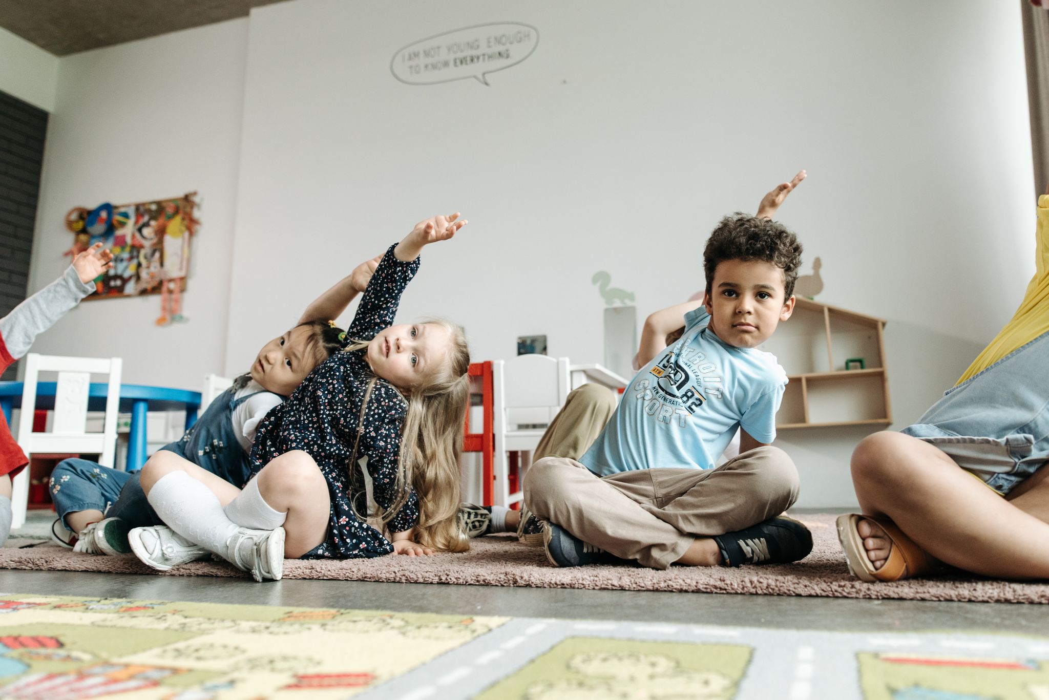 Children in a diverse classroom raising hands during a group activity indoors.